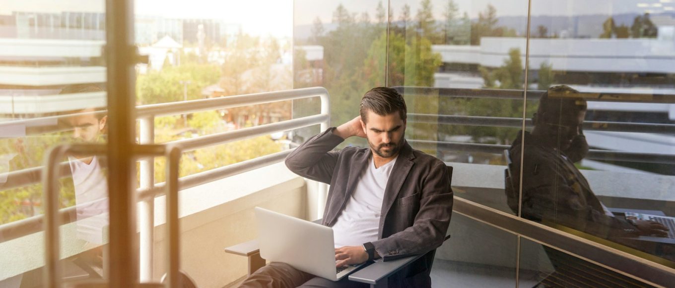 man sitting on gray arm chair using silver laptop computer on building balcony at daytime